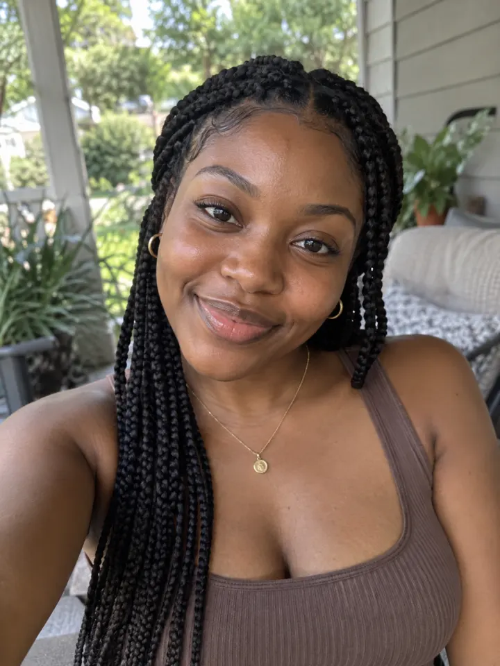 A selfie of a woman with long braids on a porch, head and shoulders in frame, smiling at the camera